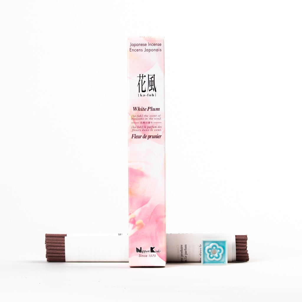 The Ka-Fuh White Plum Incense box stands upright, featuring soft pink floral designs with English and Japanese text. It’s flanked by two incense stick stacks and includes a small turquoise floral ceramic holder for a pure scent experience.