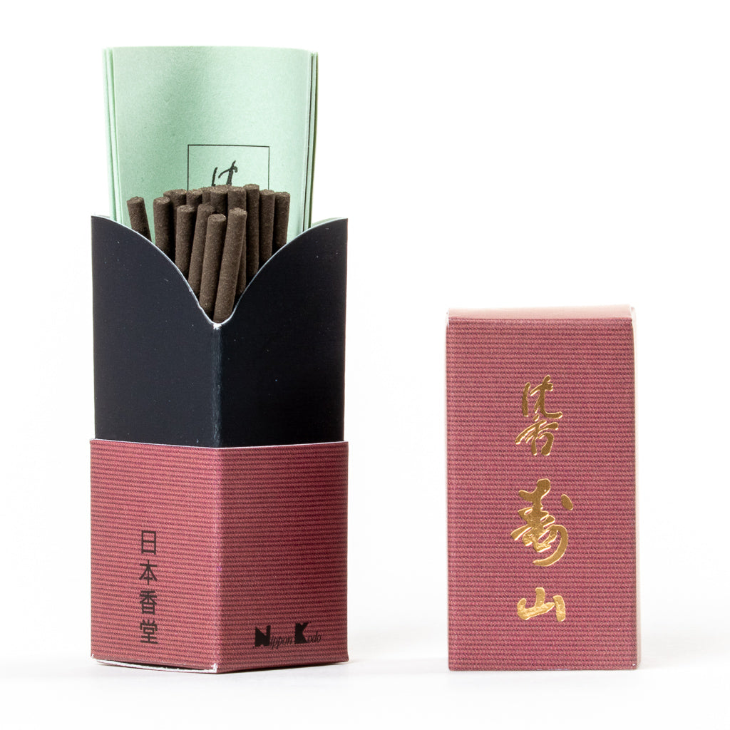 An open box of Jinkoh Juzan Aloeswood Japanese Incense, containing black aloeswood sticks and a green booklet, sits beside a closed box with gold kanji on its reddish cover. The neatly arranged incense invites tranquility and reflection.