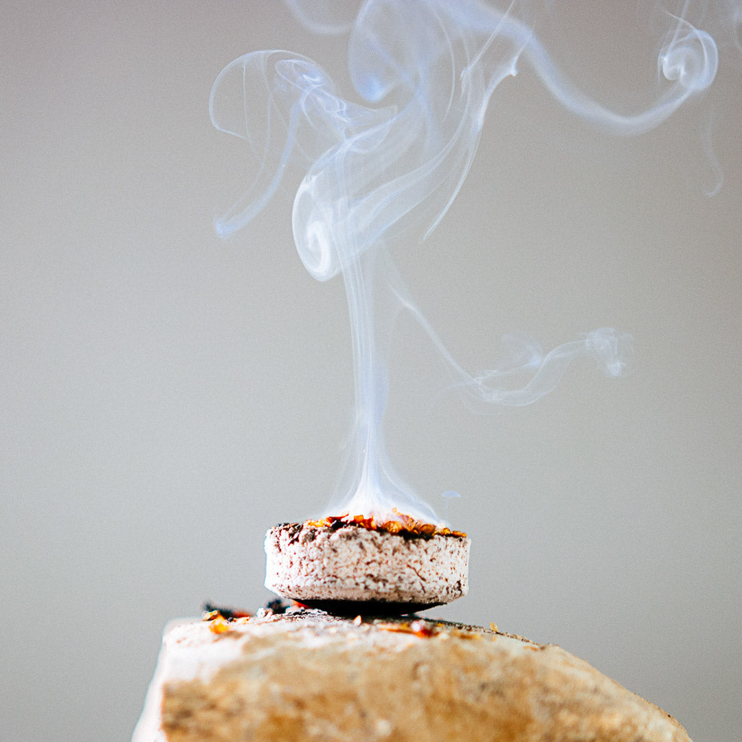 An Incense Charcoal burner rests on a stone, releasing a swirl of fragrant smoke that twists delicately upward against a plain backdrop.