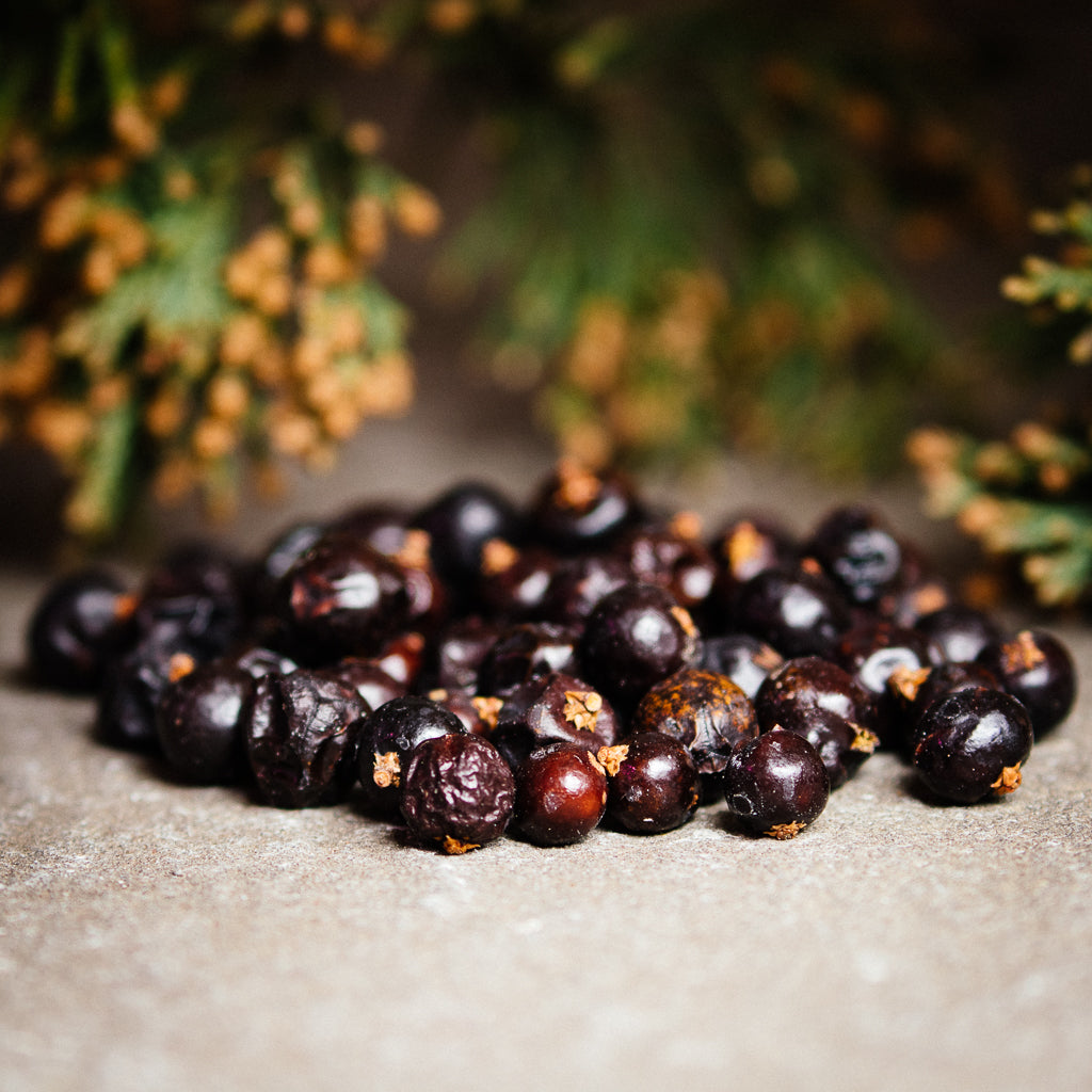 A close-up of organic juniper berries piled on a textured surface, evoking spiritual purification, with green, blurred foliage in the background.