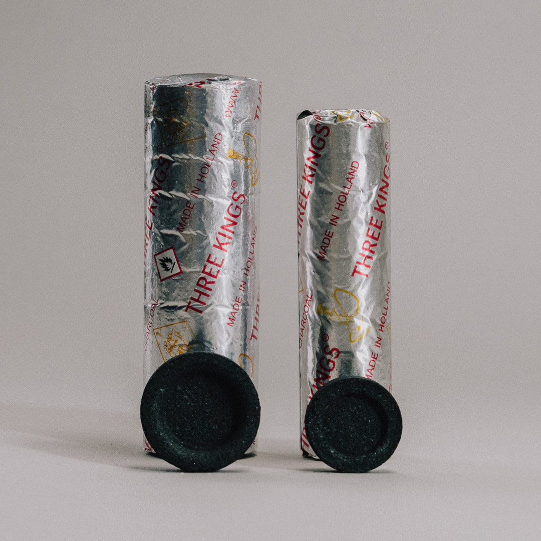 Two rolls of wrapped Incense Charcoal with shiny red text and gold crown designs stand upright against a neutral background. Two individual quick-light charcoals are placed in front, ready for purification ceremonies or burning incense.