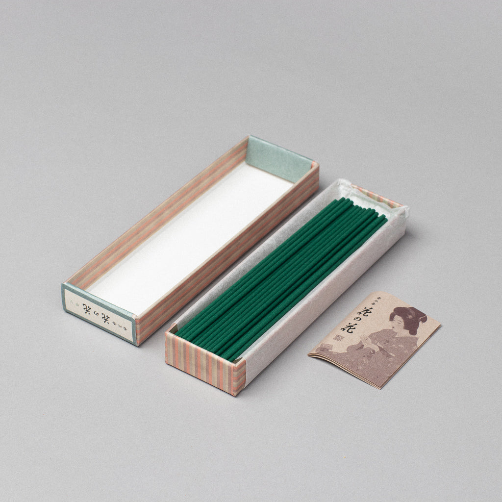 A box of Hana-no-hana Floral Japanese Incense lies open with striped edges, beside its lid and a small brown booklet on a gray background, evoking timeless elegance.
