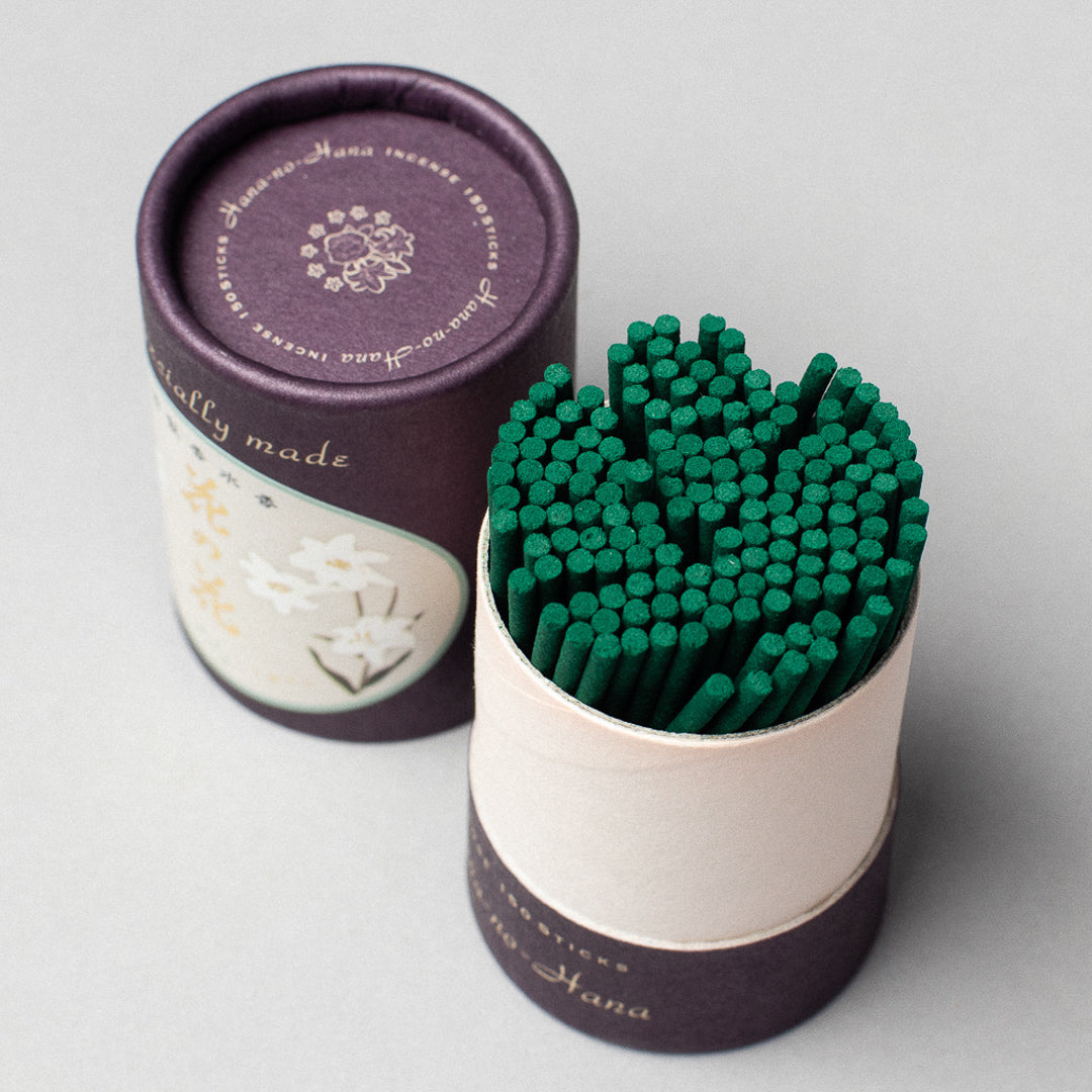 A purple and cream cylindrical container with floral designs, lid beside it, holds green incense sticks. The text reads naturally made. Infused with Hana-no-Hana, this incense named Hana-no-hana Premium Floral Japanese Incense adds elegance to your space.