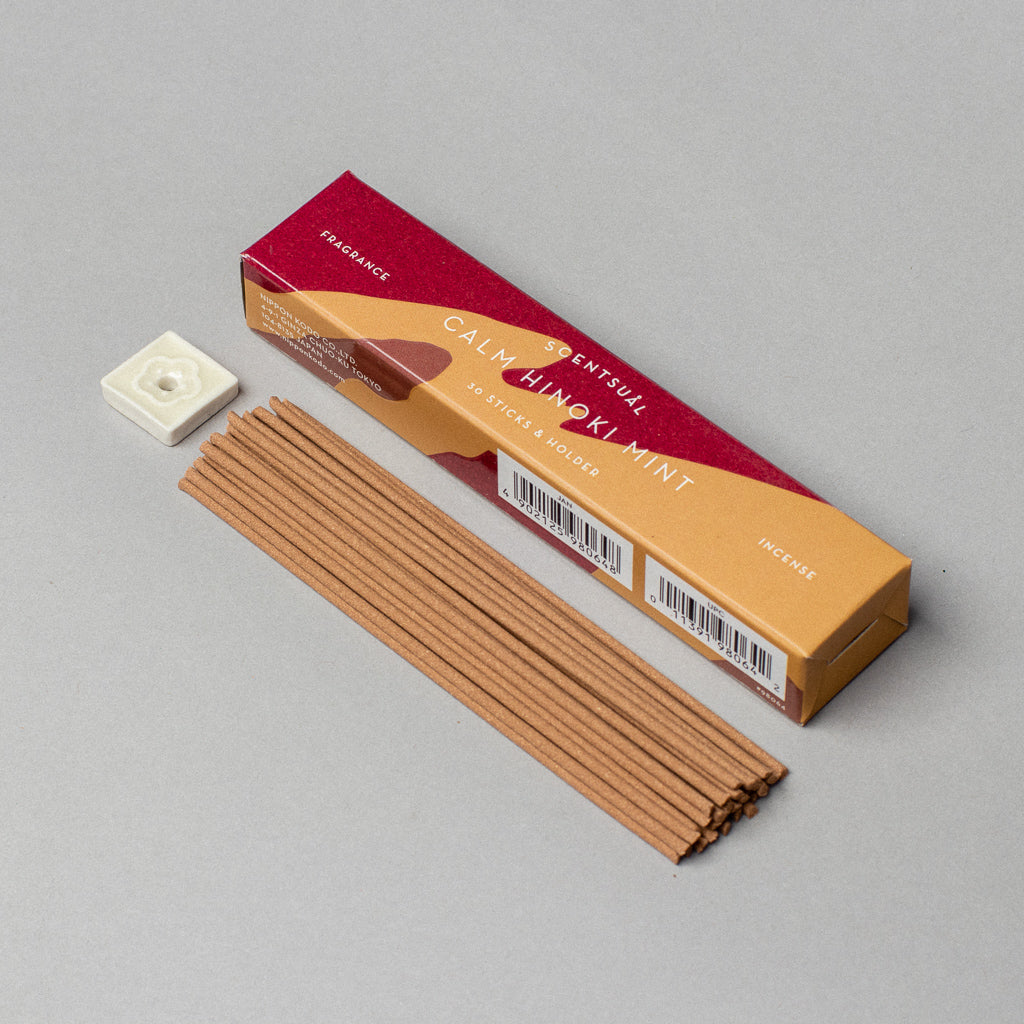 An open Scentsual Japanese Incense box reveals tan sticks beside a small white floral holder. The packaging, with minimalist red and orange designs, white text, and barcodes, contains natural incense for a fresh fragrance experience.