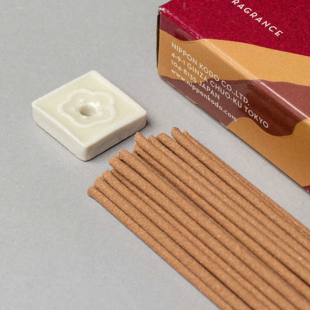 A box of Scentsual Japanese Incense sits next to a small ceramic holder with a floral design. The simple packaging shows the companys name and address in Tokyo, Japan. The brown incense sticks offer a clean fragrance, all neatly arranged on a gray surface.