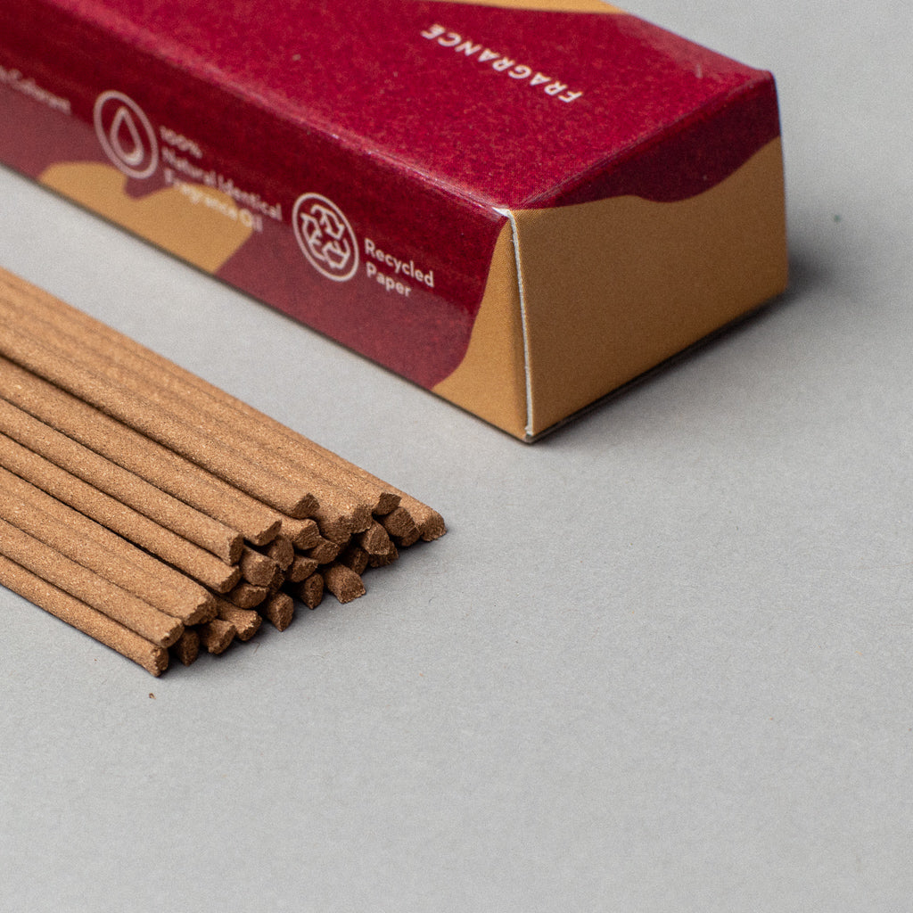 Brown incense sticks are partially pulled from a red and brown Scentsual Japanese Incense box labeled FRAGRANCE. Icons indicate natural incense and recycled paper origins, with minimal packaging on a neutral gray background that highlights its clean fragrance.