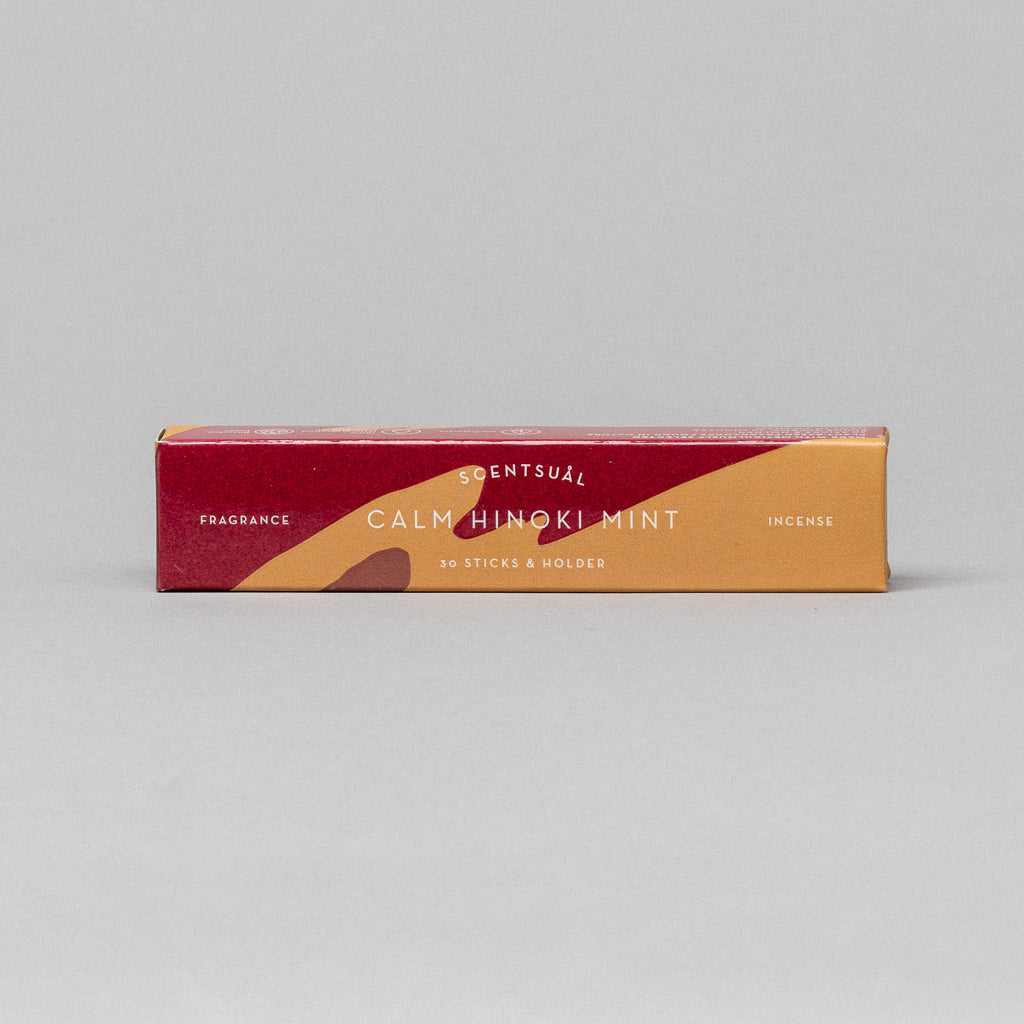 The Scentsual Calm Hinoki Mint incense box features clean, minimal packaging with a red, orange, and yellow design. It includes text specifying 30 sticks & holder and Fragrance Incense. This elegant package rests against a plain gray background.