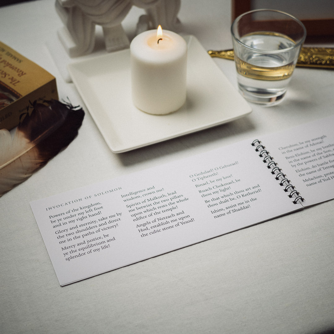 A spiral-bound Daily Prayer Book open to a page on Glorian invocations is elegantly arranged with a lit white candle on a square dish, a glass of water, feather, and decorative box, all atop a white tablecloth.