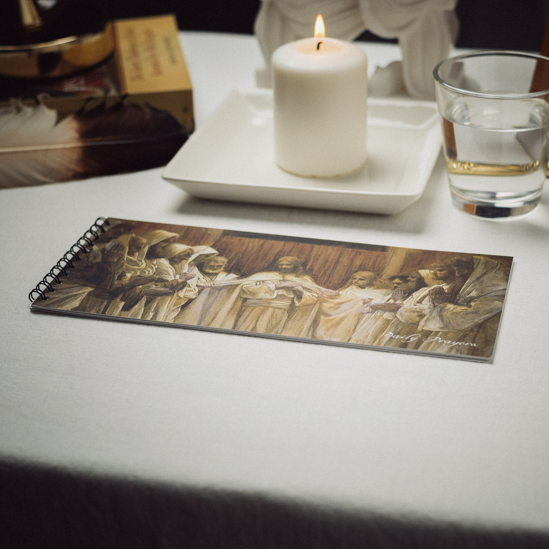 The Daily Prayer Book, spiral-bound and illustrated with people in robes performing invocations, is placed on a white tablecloth. A lit white candle and a glass of water beside it enhance the serene setting reminiscent of ancient Conjurations.