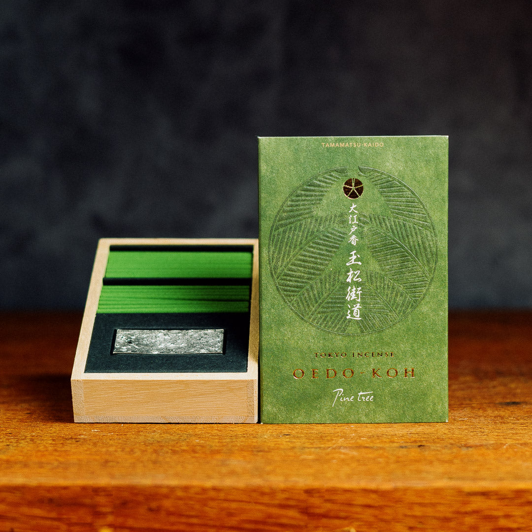 An open box of Oedo-Koh Pine Tree Incense sits on a wooden surface. The green packaging features white Japanese text and a leaf design. Inside is a black and gray incense holder, arranged elegantly against a textured gray wall.