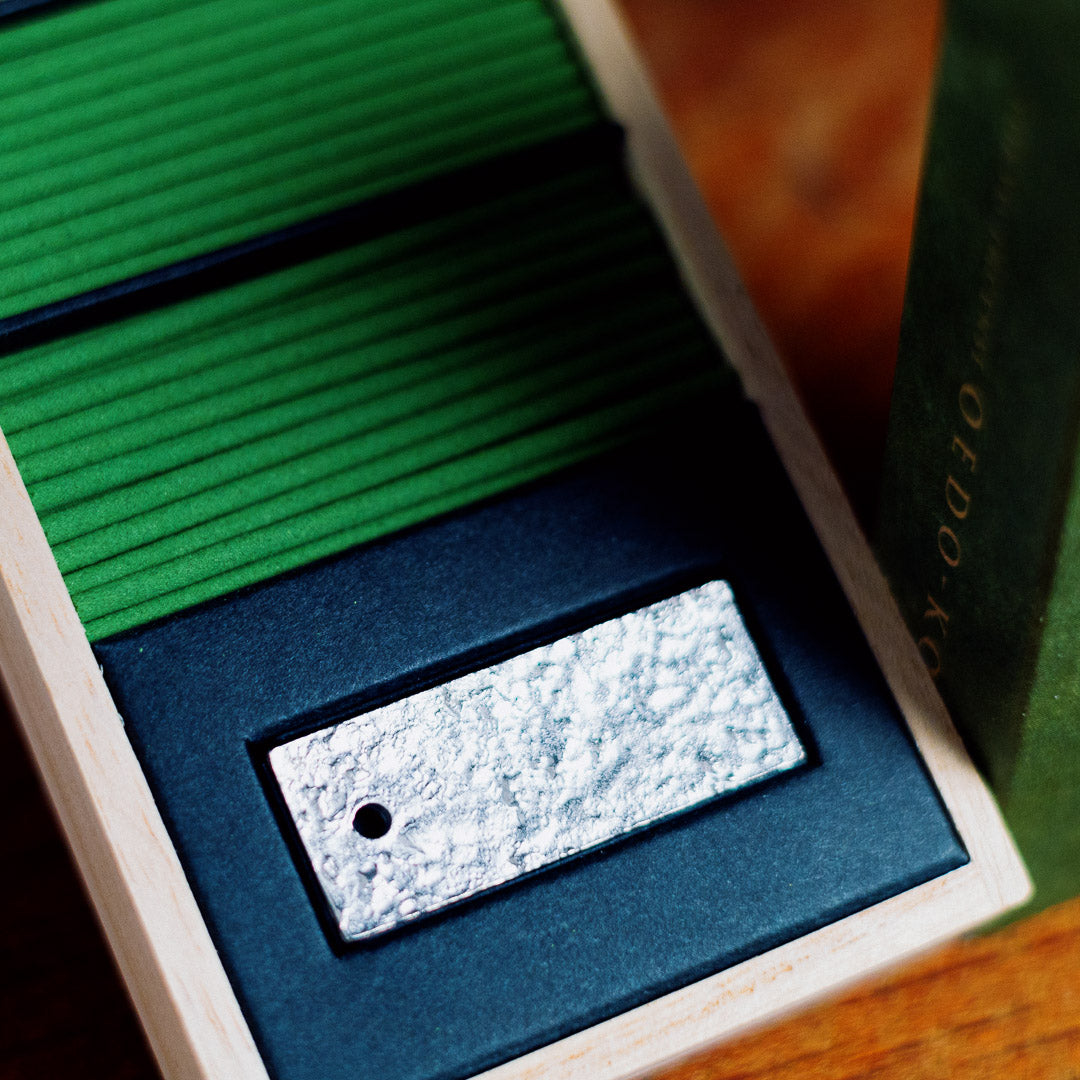 A textured silver rectangle with a small hole is set against layered green and black materials, evoking the serene aroma of Oedo-Koh Pine Tree Incense.