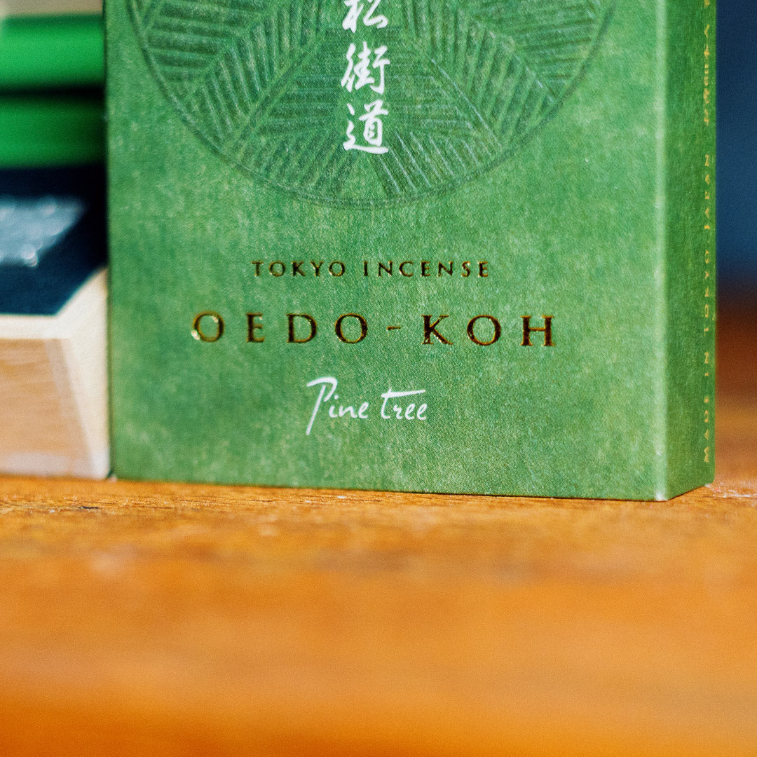 Close-up of a green Oedo-Koh Pine Tree Incense box labeled with Japanese characters. The blurred background features wooden surfaces and parts of other boxes, enhancing the elegance of this exquisite Japanese incense.