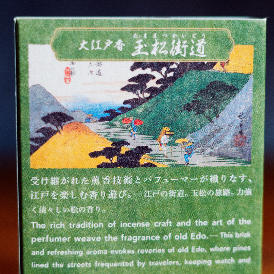 A close-up of the Oedo-Koh Pine Tree Incense gift box features traditional Japanese artwork of a rural landscape with travelers. Below the vibrant scene, Japanese and English text celebrates the rich tradition of crafting pine incense, evoking timeless fragrances of old Edo.