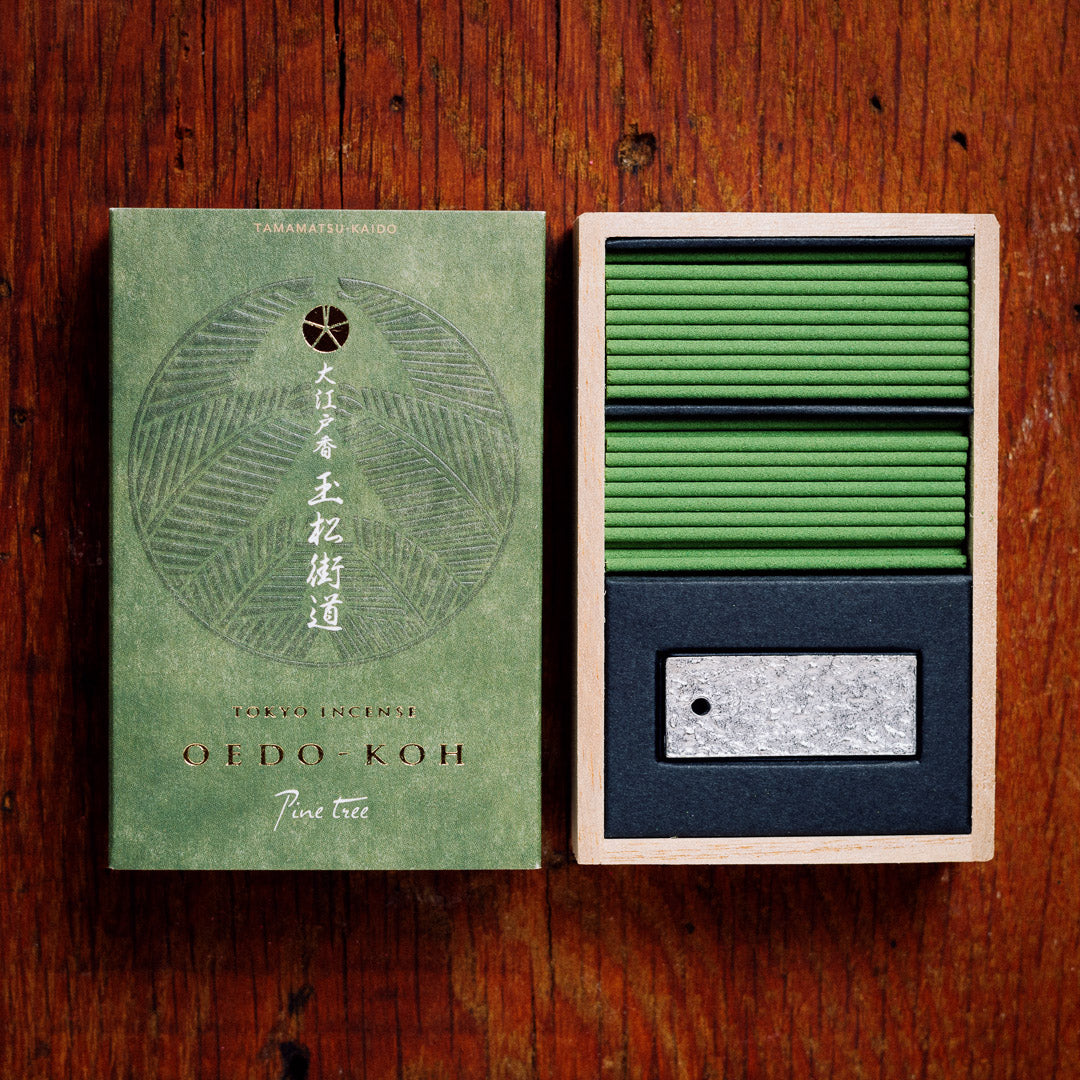 An open box of Oedo-Koh Pine Tree Incense rests on a wooden surface, revealing neatly stacked green sticks. The elegant gift box lid is adorned with graceful Japanese script and decorative designs.