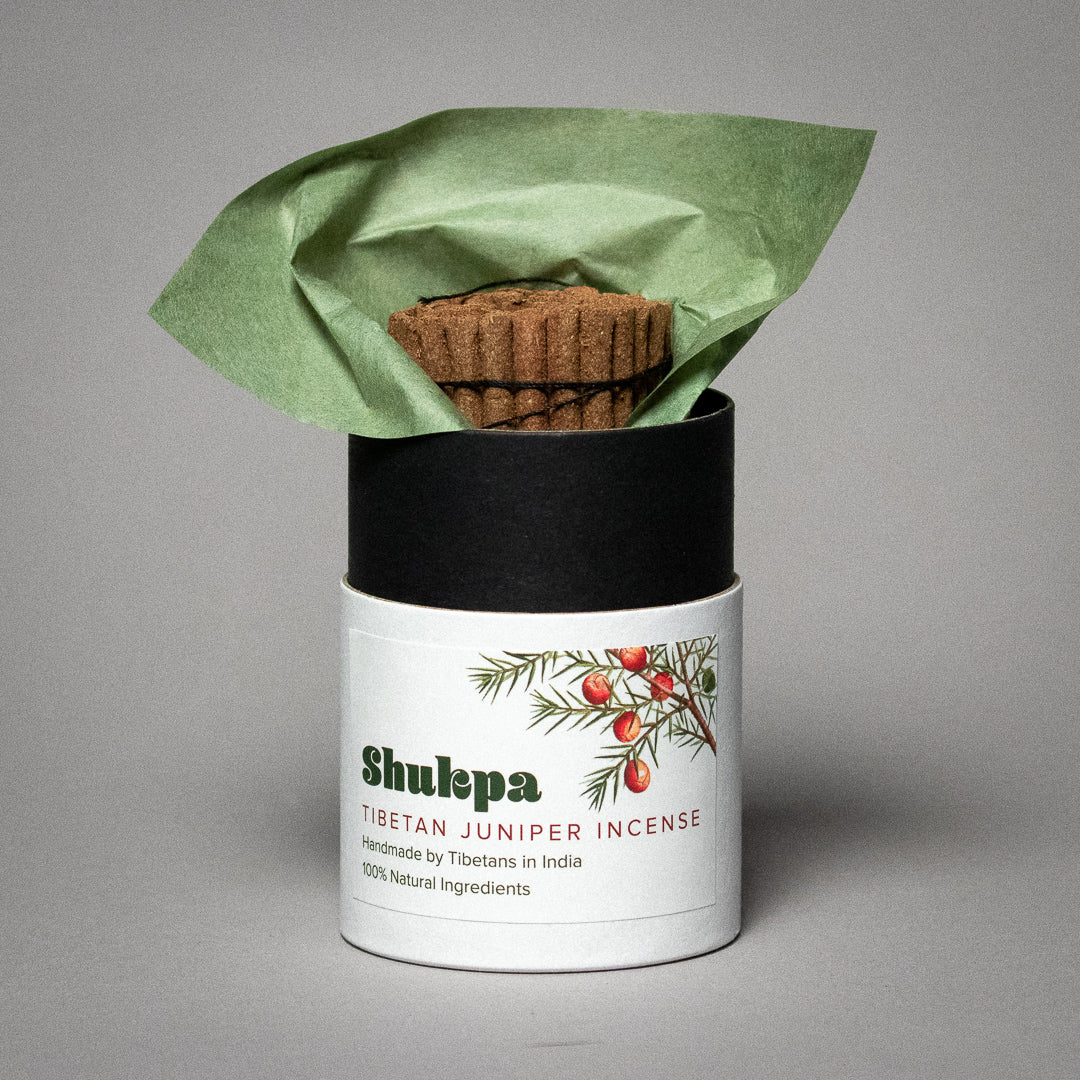 A partially open package of Shukpa Handcrafted Tibetan Juniper Incense reveals incense sticks wrapped in green paper. The label, featuring a branch with red berries, highlights its natural ingredients and authentic Tibetan craftsmanship.