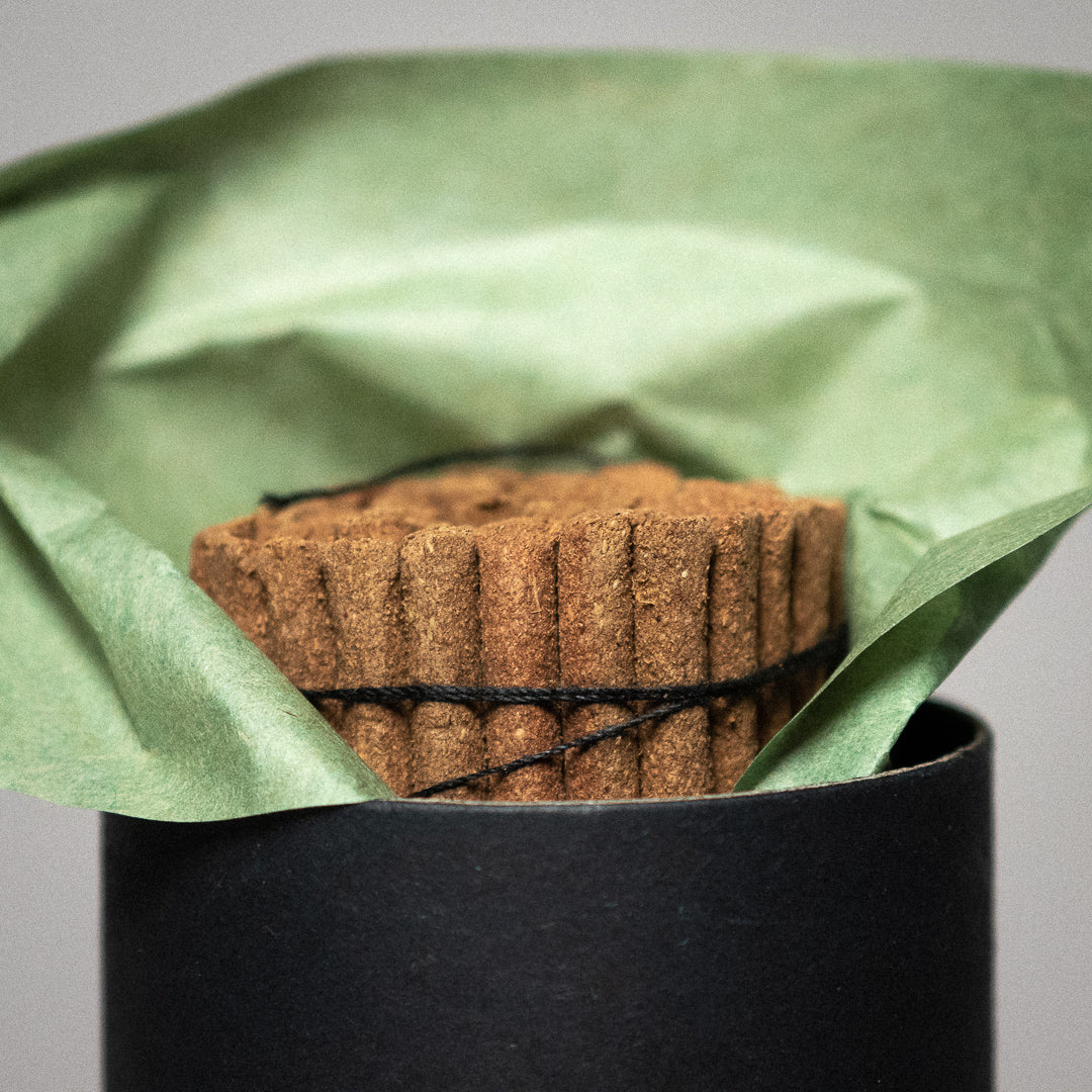 A handcrafted Shukpa Tibetan Juniper Incense, wrapped in black string, emerges from a dark cylindrical container. It rests on green paper against a soft gray background, showcasing its round and textured brown design.