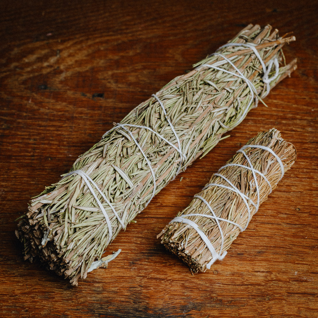 On a wooden surface lie two bundles of dried herbs tied with white string: the larger, elongated and the smaller, shorter. These light brown Rosemary Smudge Sticks with green hints evoke traditional medicines essence, reminiscent of rosemary incense.