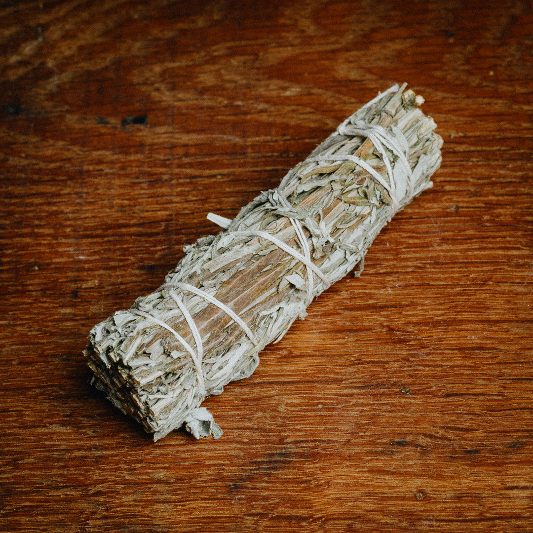 A Black Sage (Mugwort) Smudge Stick, neatly tied with string, sits on a wooden surface, prepared for cleansing rituals.