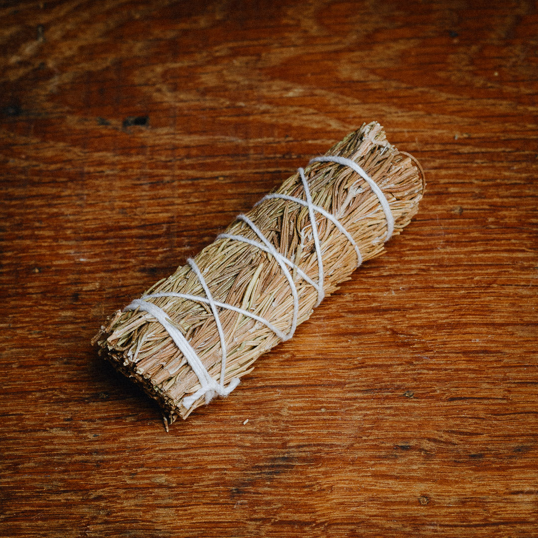 A Rosemary Smudge Stick, similar to traditional medicine, wrapped with white string rests on a wooden surface.