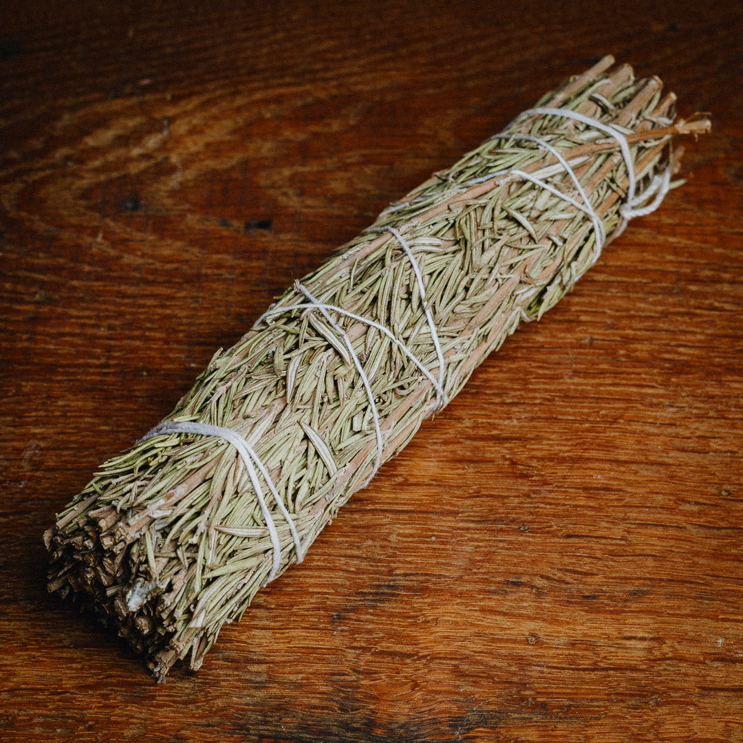A Rosemary Smudge Stick, bound with white string, rests on a wooden surface. The herbs, evocative of traditional medicine, are neatly tied to highlight their natural texture and earthy color.