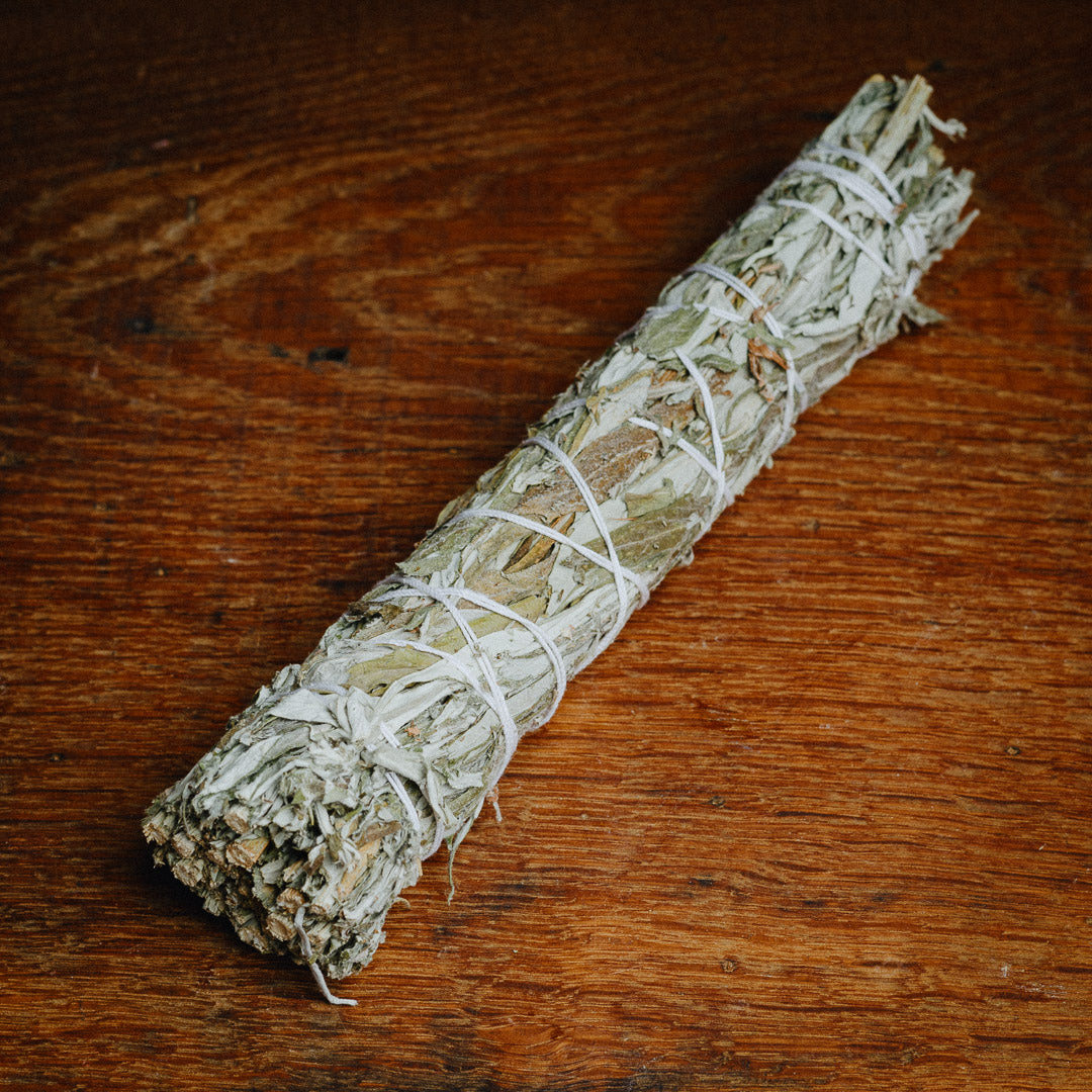A Black Sage (Mugwort) Smudge Stick, tightly bundled and tied with white string, rests on a wooden surface. The aromatic dried leaves are packed and wrapped, perfect for smudging or cleansing rituals.
