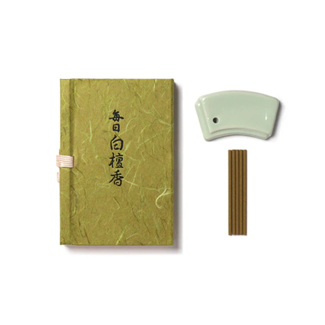 The Mainichi Byakudan Premium Sandalwood Japanese Incense set includes a green textured box with elegant calligraphy, a small white ceramic holder, and several sandalwood sticks, perfect for enhancing your meditation fragrance routine.