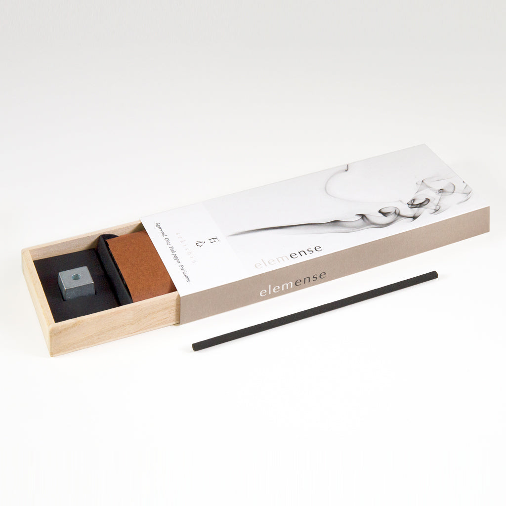 An open box of Elemense Sekishin Japanese Incense is shown on a white surface. It includes a black incense stick, metal holder, and book. The minimalist packaging, crafted from natural aromatic materials, embodies elegance inspired by the Five Elements Theory.