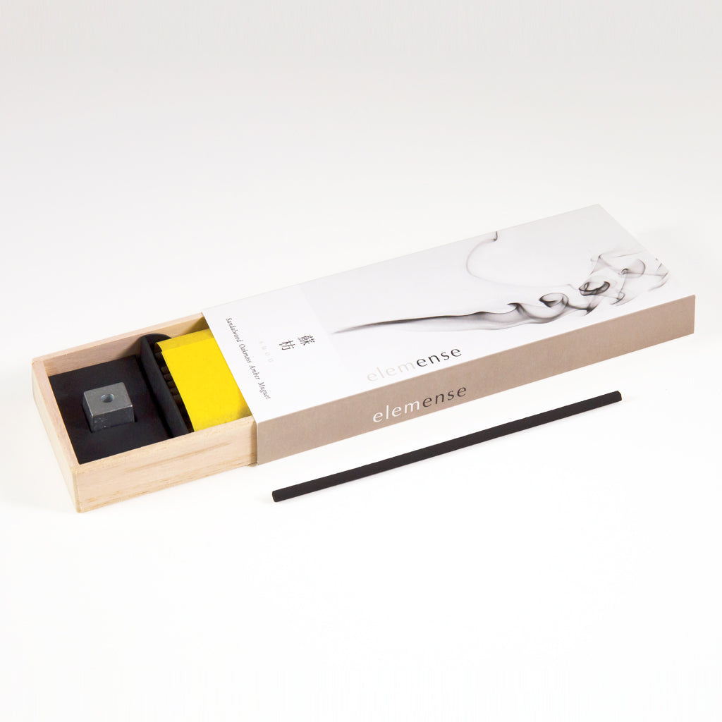 An open wooden box contains the minimalist design of the Elemense Suou Japanese Incense set, which includes a black incense stick, yellow felt mat, and stone holder. The lids smoke design reflects elegant natural aroma materials inspired by the Five Elements Theory.